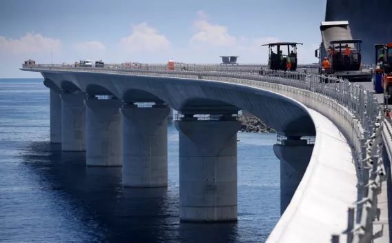 Enrobé en site maritime sur viaduc côtier courbe, finisseuse alimentée et compacteurs à l’ouvrage, équipe en orange sécurisée au-dessus de l’eau.
