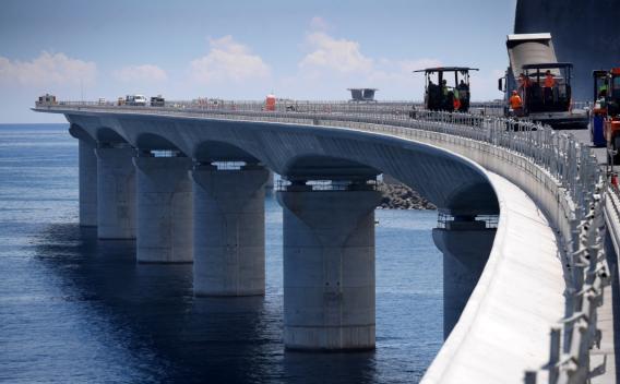 Enrobé en site maritime sur viaduc côtier courbe, finisseuse alimentée et compacteurs à l’ouvrage, équipe en orange sécurisée au-dessus de l’eau.