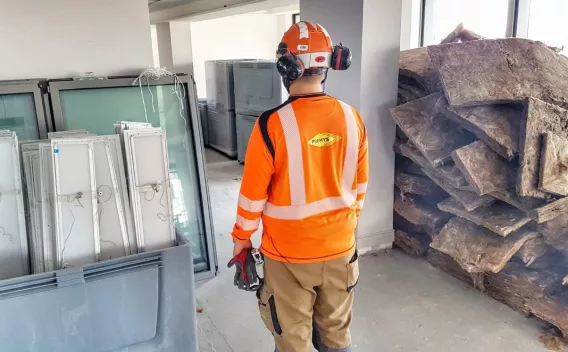 Ouvrier en tenue de chantier orange (gilet, casque, protections) debout dans un local de chantier intérieur, entouré de menuiseries et d'isolant empilés.