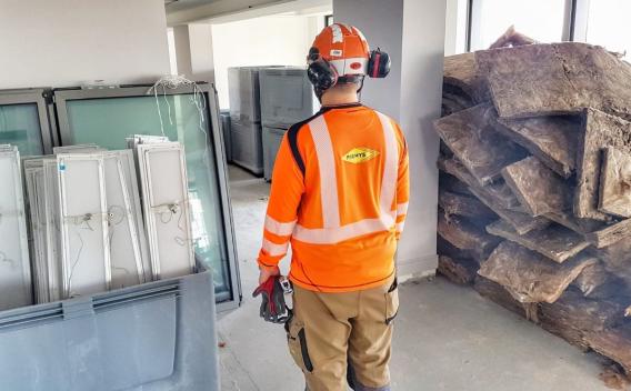 Ouvrier en tenue de chantier orange (gilet, casque, protections) debout dans un local de chantier intérieur, entouré de menuiseries et d'isolant empilés.