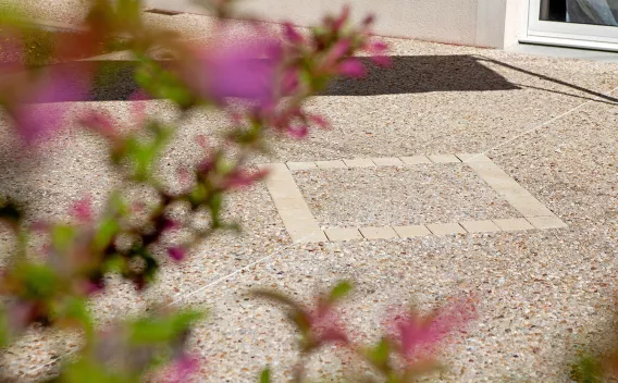 Une terrasse texturée en gravillons, encadrée en avant-plan par des fleurs roses floues et ponctuée d’un carré de pavés clairs au centre.