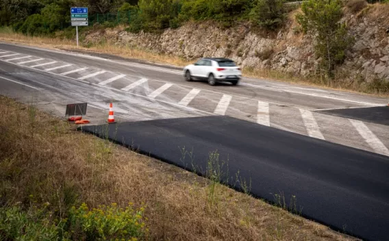Bande de chaussée fraîchement rechargée en enrobé noir le long d’un échangeur, avec cône de signalisation et véhicule passant à proximité d’un talus boisé.