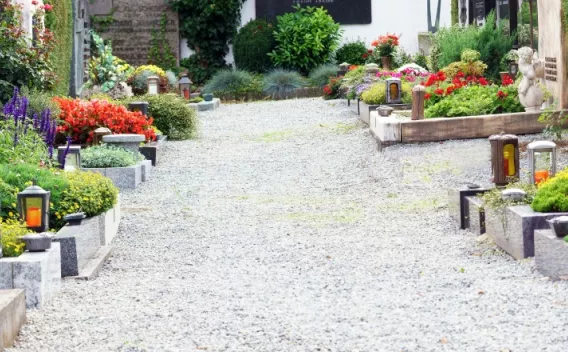 Allée de cimetière avec tombes bordées de massifs fleuris et lanternettes, sol en gravier et végétation soignée de part et d’autre.