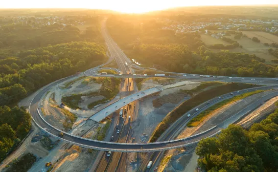 Échangeur autoroutier en travaux au coucher du soleil : vue aérienne d’un chantier de reconfiguration avec bretelles neuves, remblais en cours, circulation maintenue sur les voies centrales.