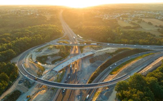 Échangeur autoroutier en travaux au coucher du soleil : vue aérienne d’un chantier de reconfiguration avec bretelles neuves, remblais en cours, circulation maintenue sur les voies centrales.