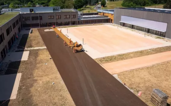 Cour d’établissement scolaire en chantier : pose d’un enrobé noir sur allée centrale, engins compacts en action, zones sportives claires attenantes et bâtiments modernes en U autour.