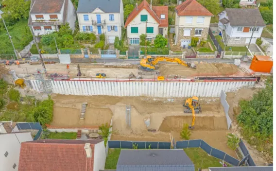 Génie civil par Colas : Vue aérienne d’un chantier de soutènement en zone résidentielle montrant palplanches installées, terrassement en cours et engins de chantier avec signalisation et stockage sur place.