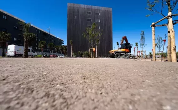 Un engin de chantier et des aménagements paysagers en cours sur une place urbaine devant un bâtiment moderne, illustrant les travaux d'embellissement et d'accessibilité.