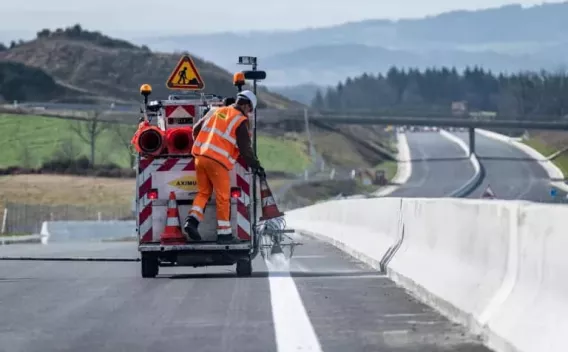 Aménagement de la RN88 par Aximum : Marquage de chaussée sur autoroute neuve : équipe en EPI appliquant une ligne blanche continue le long d’un muret central, circulation encore fermée en arrière-plan.