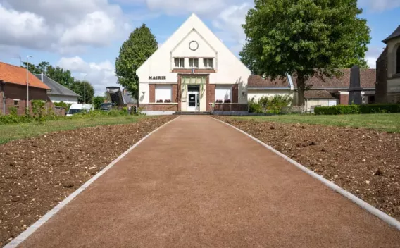 Un chemin d’accès piéton en perspective menant à une petite mairie blanche, surface de sol couleur terre cuite uniforme, bordures en béton, massifs de terre fraîchement préparés de chaque côté, arbres et bâtiments en arrière-plan sous un ciel partiellement nuageux.