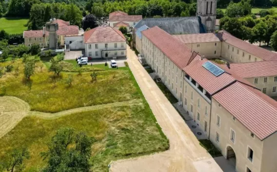 Vue aérienne d’un site patrimonial réhabilité : longue allée claire en matériaux stabilisés longeant un bâtiment historique à toits en tuiles, espaces verts fauchés à gauche, clocher et arbres en arrière-plan, quelques véhicules légers stationnés près des bâtiments.