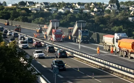 Chantier de réfection sur pont autoroutier : rabotage et mise en enrobé en voie neutralisée, file de camions bennes alimentant finisseurs, compacteurs en chaîne, circulation maintenue sur voies adjacentes et personnels en EPI orange.