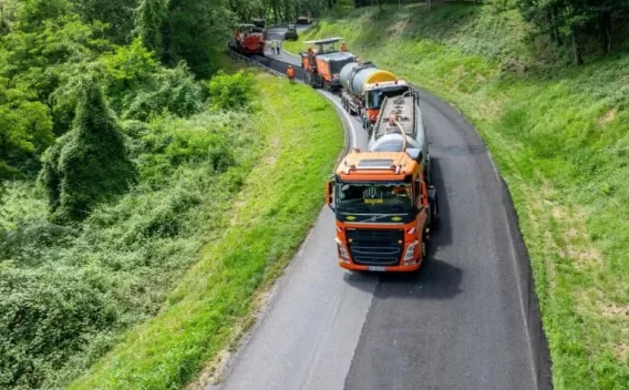 Convoi routier d’enrobés orange en train de rénover une route en courbe, plusieurs engins et camion-citerne alignés, surface fraîchement posée et bande centrale visible, entouré d’un talus végétalisé.