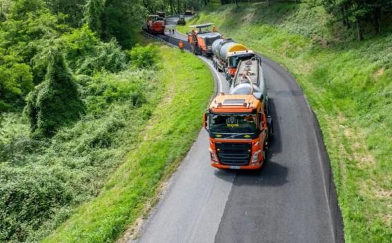 Convoi routier d’enrobés orange en train de rénover une route en courbe, plusieurs engins et camion-citerne alignés, surface fraîchement posée et bande centrale visible, entouré d’un talus végétalisé.