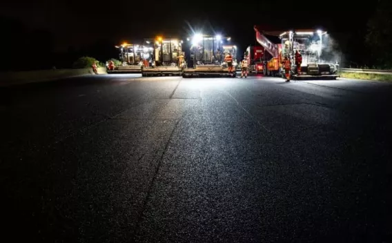 Chantier de pose d’enrobé de nuit avec une ligne d’engins éclairés et opérateurs en EPI assurant une intervention sécurisée pour la rénovation d’une chaussée.