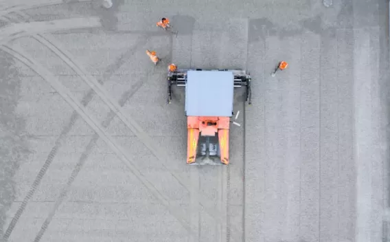 Un largage de grave non compactée vu en vue aérienne: engin de répartition orange au centre et quatre ouvriers en tenues orange, traces de pneus et bandes de matériau sur le tablier.