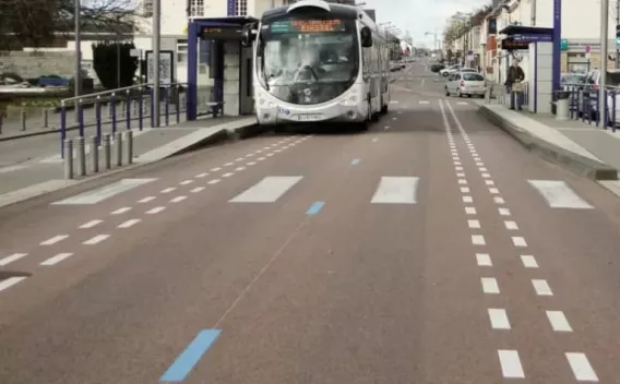 Axe de BHNS en centre‑ville : chaussée en enrobé rouge, marquages blancs et bleus dédiés, quais bus surélevés avec garde‑corps, mobilier urbain violet et circulation encadrée par bordures béton.