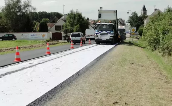 Équipe en tenues haute visibilité réalisant la mise en œuvre et la finition d’un revêtement piétonnier/voirie sur trottoir et chaussée, avec engins (dépose/pavage/compactage), balisage et stockage de matériaux sur site.