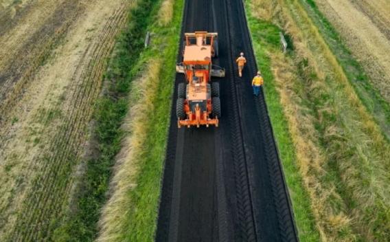 Raboteuse sur voie rurale encadrée de champs, deux opérateurs à pied supervisant une passe de fraisage.