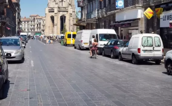 Rue ensoleillée avec voitures garées, une personne à vélo au centre et une grande porte monumentale en fond.