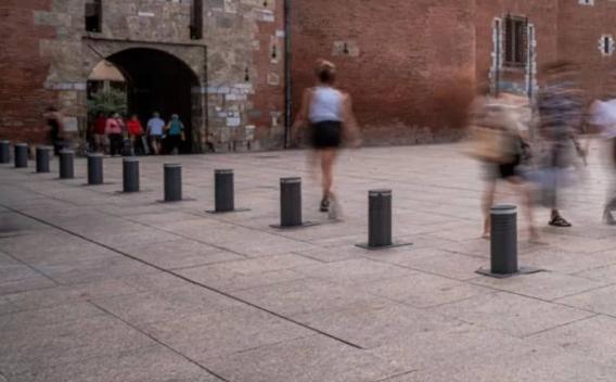 Place urbaine piétonne pavée en dalles claires, bordée de bornes escamotables alignées et d'un porche en briques au fond ; plusieurs passants flous en mouvement.