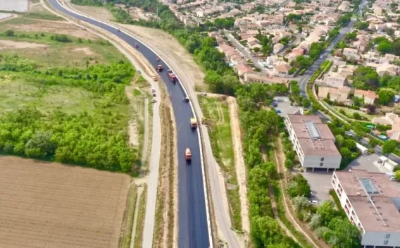Mise en œuvre et finition d’enrobé sur voirie en milieu périurbain : équipes en tenues haute visibilité et engins d’enrobage/compactage opérant sur une chaussée sinueuse, accotements préparés et zone de chantier balisée.