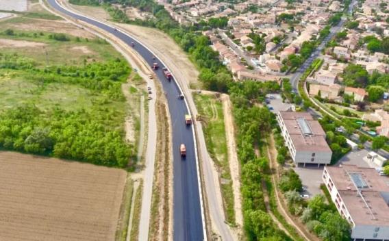 Mise en œuvre et finition d’enrobé sur voirie en milieu périurbain : équipes en tenues haute visibilité et engins d’enrobage/compactage opérant sur une chaussée sinueuse, accotements préparés et zone de chantier balisée.
