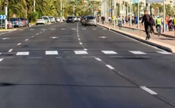 Boulevard urbain littoral : enrobé noir récent, passages piétons et pointillés centraux bien contrastés, bordures granit et promenade plantée de palmiers à droite, trafic diffus en arrière‑plan.
