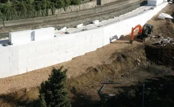 Mur de soutènement blanc en blocs isolants le long d’un talus, pelle mécanique à l’œuvre et terrassement en cours.
