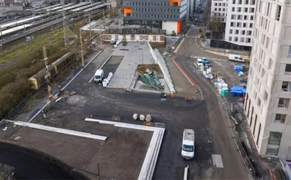 Travaux d'aménagement urbain en cours avec voirie asphaltée, îlots minéraux, engins et véhicules de chantier, et coordination logistique autour d'un nouveau pôle urbain.