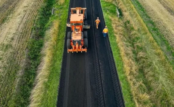 Deux opérateurs en tenue de chantier et un engin règlent la pose et le compactage d’une couche d’enrobé sur une voie rurale, assurant contrôle de la qualité et sécurité.