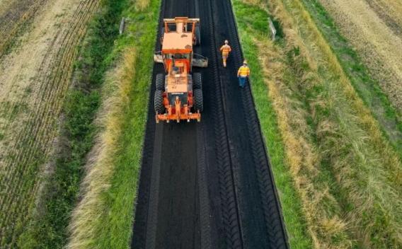Deux opérateurs en tenue de chantier et un engin règlent la pose et le compactage d’une couche d’enrobé sur une voie rurale, assurant contrôle de la qualité et sécurité.