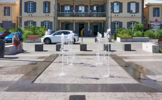Place publique pavée avec jets d'eau décoratifs en premier plan, mobilier urbain et bâtiment administratif en arrière‑plan.