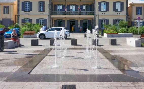 Place publique pavée avec jets d'eau décoratifs en premier plan, mobilier urbain et bâtiment administratif en arrière‑plan.