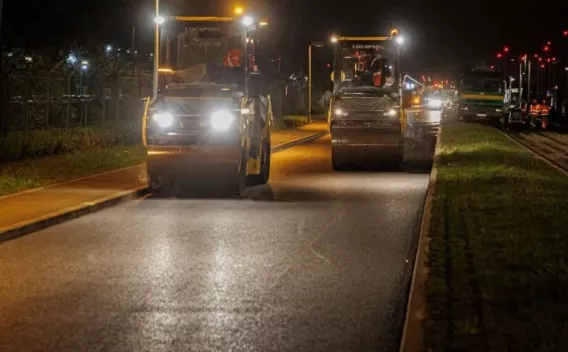 Compactage nocturne de chaussée : deux rouleaux tandem illuminés progressent sur une voirie urbaine, encadrés par trottoirs et bande engazonnée, illustrant travaux de finition et continuité des opérations de nuit.