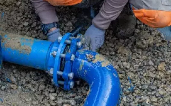 Installation canalisation eau par Colas : Raccordement de canalisation eau potable : agent en EPI serrant un collier de bride sur tuyau bleu dans une tranchée gravillonnée.