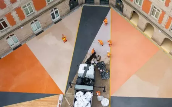 Cour intérieure en rénovation avec revêtements colorés géométriques : larges triangles d’enrobés ou bétons décoratifs (orange, beige, gris foncé) rayonnant depuis un angle, opérateurs en tenue haute visibilité à l’œuvre autour d’un camion et d’engins légers.