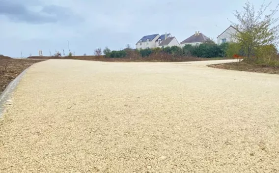 Allée en enrobé drainant clair au ras du sol, légère montée vers un lotissement naissant sous un ciel nuageux.