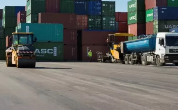 Chantier d’enrobé sur aire logistique : pose de revêtement bitumineux à l’aide d’une finisseuse alimentée par un camion benne, rouleau compacteur en manœuvre pour assurer la compaction. Zone dégagée entourée de conteneurs, procédure de mise en œuvre conforme aux opérations de revêtement lourd (rabotage/pose, alimentation continue, compactage).