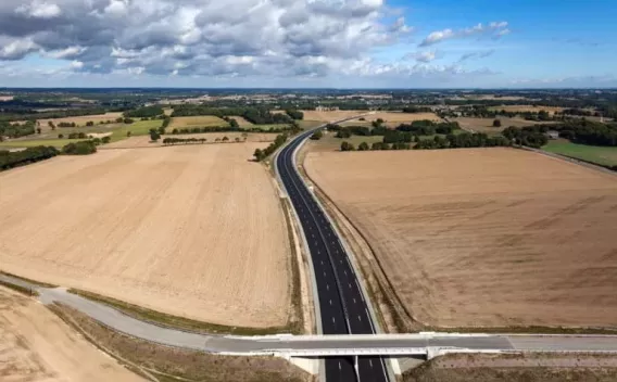 Vue aérienne d’une route neuve traversant des zones agricoles, avec pont et infrastructure moderne.