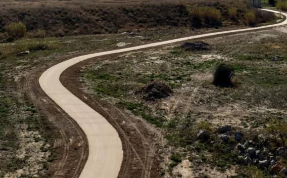 Cheminement neuf en enrobé clair serpentant dans un terrain naturel semi-aride : ruban continu aux bords nets, talus et végétation clairsemée autour, tracé sinueux s’éloignant vers l’horizon.
