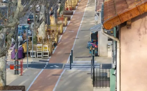 Un quai piéton/cyclable en bordure de rue commerçante, bandes blanches délimitant voies, mobilier et terrasses rangés, perspective en légère plongée.
