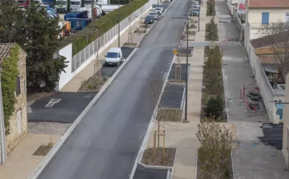 Rue urbaine rénovée vue en contre‑plongée : chaussée et trottoirs neufs bordés d'îlots plantés, places de stationnement et aménagements piétons.