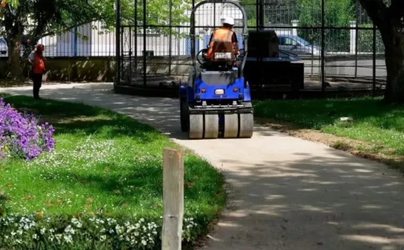 Compactage d’une allée piétonne en stabilisé dans un parc, petit rouleau tandem en action sous la supervision d’un opérateur, massifs fleuris et pelouse autour.