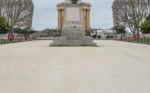 Parvis urbain en cours d’aménagement : large esplanade en grave claire devant un monument classique, zone périmétrée par des barrières de chantier, présence d’un camion-citerne et de signalisation, arbres taillés en têtard de part et d’autre.