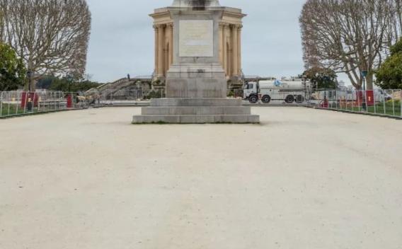 Parvis urbain en cours d’aménagement : large esplanade en grave claire devant un monument classique, zone périmétrée par des barrières de chantier, présence d’un camion-citerne et de signalisation, arbres taillés en têtard de part et d’autre.