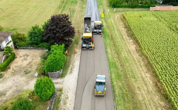 Pose d’enrobé en milieu rural : finisseuse alimentée par camion benne, suivi par un rouleau compacteur pour assurer l’homogénéité et la compaction de la couche, interventions réalisées en bande continue le long d’une parcelle agricole.