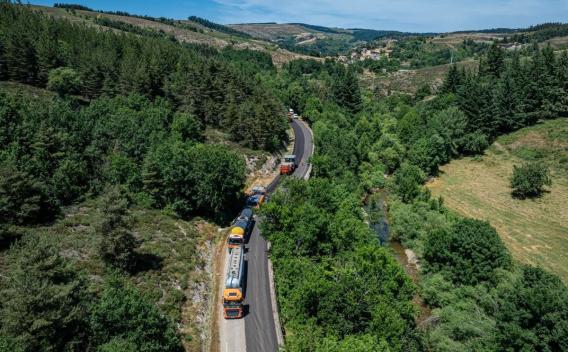 Vue aérienne d'une carrière à ciel ouvert en terrasses creusées dans un versant boisé, avec rampes d'accès hélicoïdales et zones de stockage et concassage en pied de site ; le pourtour est dominé par des prairies et massifs boisés.