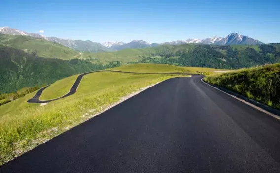 Route neuve en montagne avec enrobé noir lisse sinueux au premier plan et paysage de collines verdoyantes à l'arrière-plan.