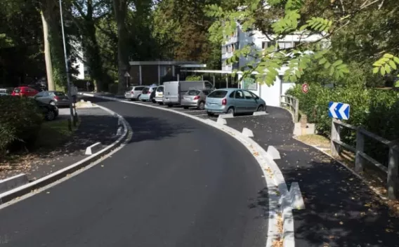 Voirie urbaine rénovée sous couvert boisé : enrobé noir lisse, caniveaux et bordures béton neufs, stationnements latéraux et giratoire à l’arrière-plan.